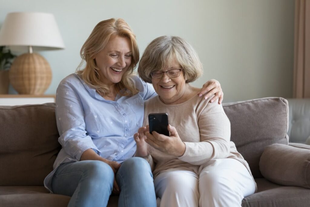 Happy nurse, caregiver helping cheerful elderly grandma woman to use online app on mobile phone